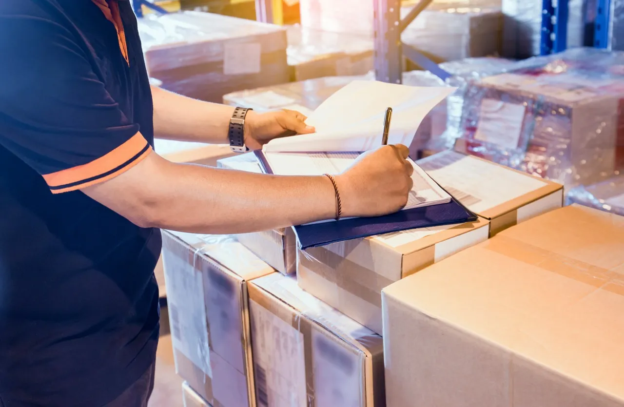 A worker checking paperwork on top of boxed cargo in a warehouse